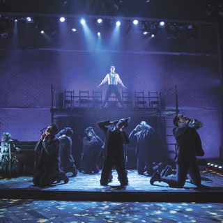 An a scene from Ӱɴý's presentation of the musical Spring Awakening, an ensemble of actors in period costume kneel while a male soloist sings under a blue spotlight on an elevated platform.
