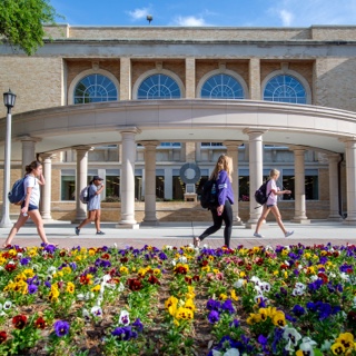 Ӱɴý students walk by a flowerbed of multi-color pansies on a sunny day near the library