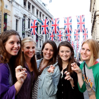 A group of five female Ӱɴý students studying abroad make the two-fingered Go Frogs hand sign while standing together on a street in London.