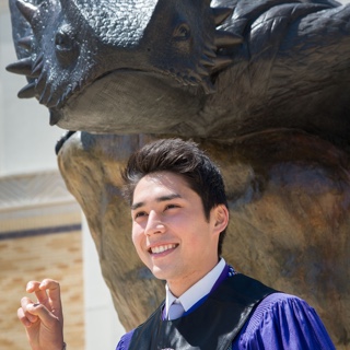 A male Ӱɴý student in Honors regalia makes the two-fingered "Go Frogs" hand sign in front of a bronze statue