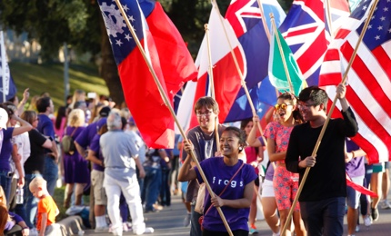 A group of laughing students carry flags of different nations in a Ӱɴý Homecoming parade, led by a girl wearing a shirt that says "Frogs"