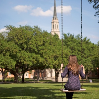 Back of female Ӱɴý student sitting in a metal swing with a cluster of trees and the Ӱɴý chapel in the background.