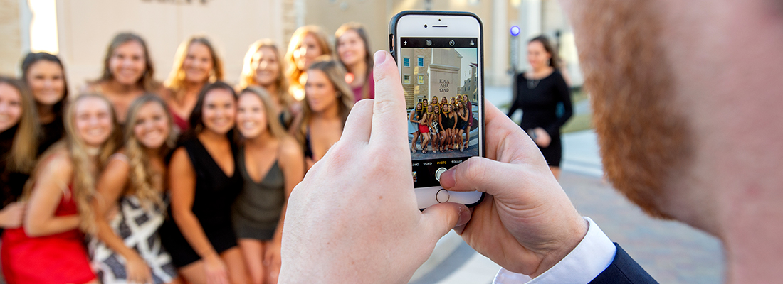 A young man uses a cell phone to photograph a group of smiling sorority girls before a party in the Greek Village at Ӱɴý.