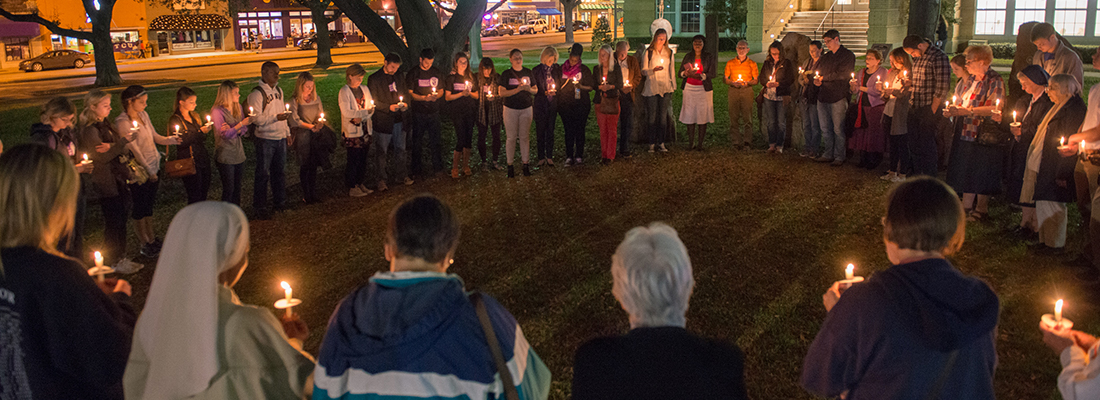 A circle of people holding candles gather for a nighttime prayer vigil on the lawn of the Ӱɴý campus.